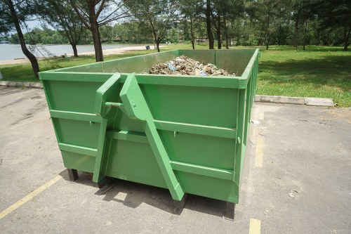 A large, green industrial skip filled with a mixture of garden and household waste, including broken branches, soil, and discarded materials, positioned on a paved parking area near a grassy park with trees and a body of water in the background. The skip is made of metal with reinforced sides and is situated adjacent to a curb, suggesting it is prepared for rubbish removal services in the Chessington area. The scene is outdoors, with natural daylight illuminating the skip and its surroundings, indicative of a typical waste collection environment managed by gardening or rubbish removal specialists such as Garden Clearance Chessington.