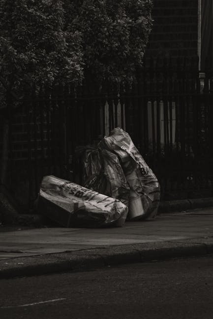 A photograph shows a pile of mixed household rubbish placed on a paved driveway or garden path, with a background of a building with brick and white siding. The pile contains several black plastic gar